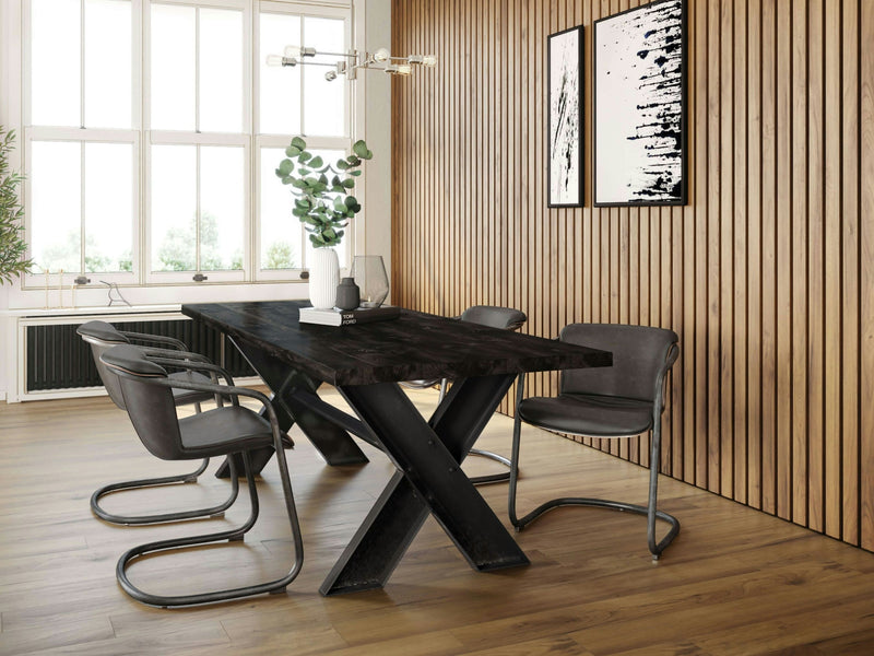 A modern dining room features the X-Base Dining Table - Charred Ember by James+James, with a wooden top and a black, V-shaped metal base, surrounded by four grey chairs. A vase with greenery sits on the table. The room has large windows, wooden panel walls, and abstract black-and-white paintings.