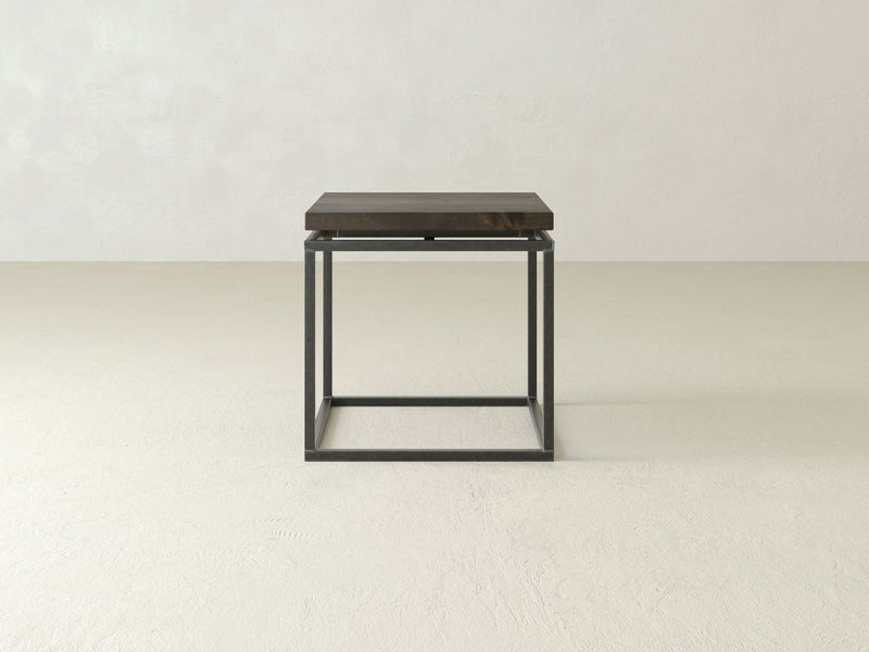 The Floating Top End Table - Deep Grey by James+James features a minimalist design with a dark wooden top and a black metal frame. Positioned in an empty, light-colored room with a textured floor, the table embodies simplicity and modernity, emphasizing clean lines and an open aesthetic.