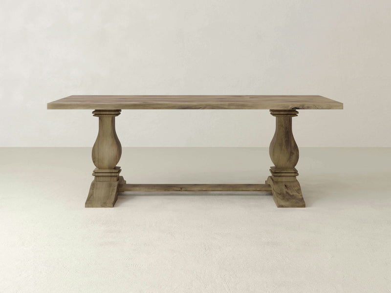 The Heirloom Dining Table - Harvest Wheat by James+James is a wooden dining table with a rectangular top and two pedestal legs, each featuring ornate carvings. The table has a rustic, unfinished appearance and is placed against a plain, light-colored background.