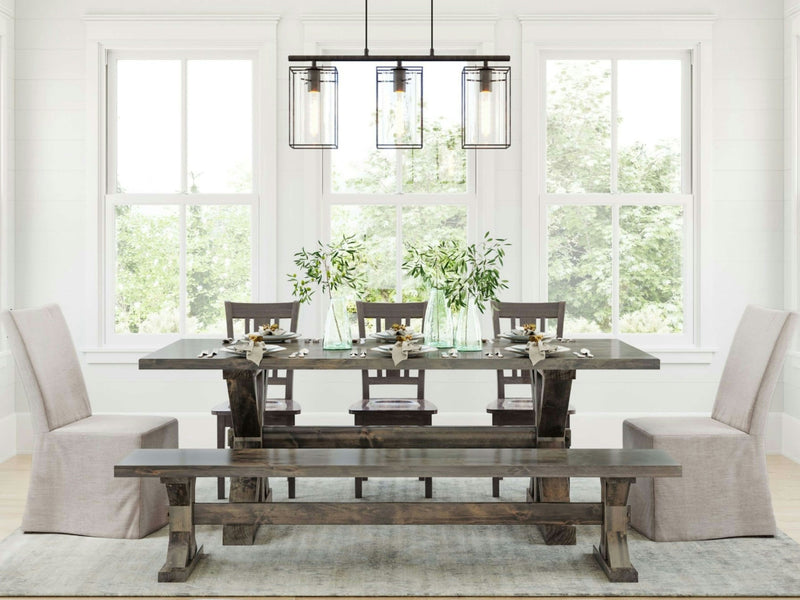 A bright dining room features a Trestle Dining Table in Deep Grey by James+James, set with plates, glasses, and greenery centerpieces. Four wooden chairs with light-colored cushions and a matching wooden bench surround the table. Large windows behind the table let in natural light, illuminating the space.