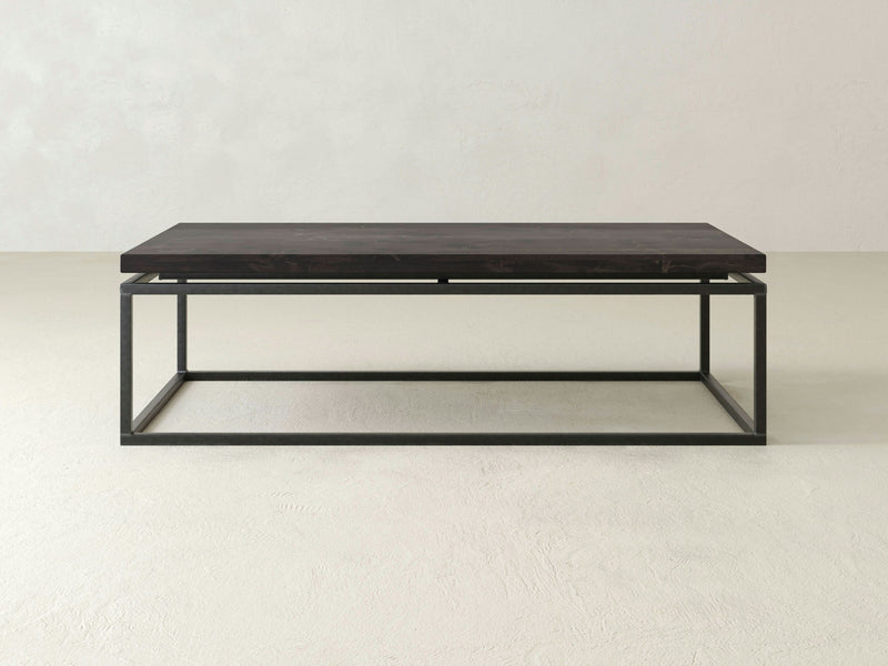 A rectangular coffee table, the "Floating Top Coffee Table - Charred Ember" by James+James, features a dark wooden top paired with a minimalist black metal frame. The table boasts a simple, modern design and is set against a plain, light-colored background.