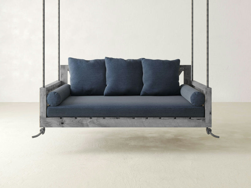 A suspended wooden sofa, featuring the Mattress Cushion and Pillows for Crib Swing from James+James—with durable, Sunbrella blue cushions and two matching bolster pillows—hangs from the ceiling by thick ropes against a plain white wall and light-colored floor, creating a minimalist and airy atmosphere.