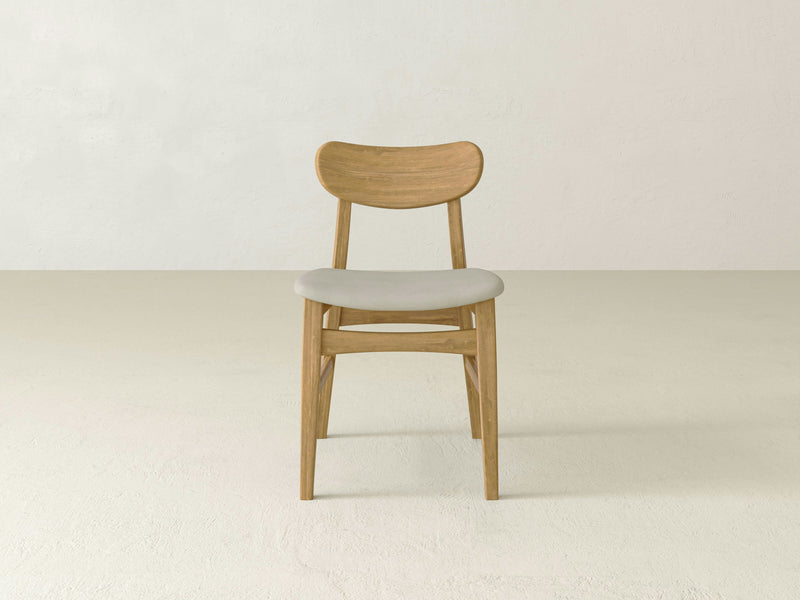 A Lynn Upholstered Dining Chair - Harvest Wheat by James+James is centered in the image. The chair features a minimalist design with a curved backrest, four simple legs, and a light beige seat cushion. The background consists of an off-white wall and floor, contributing to the clean and modern aesthetic.