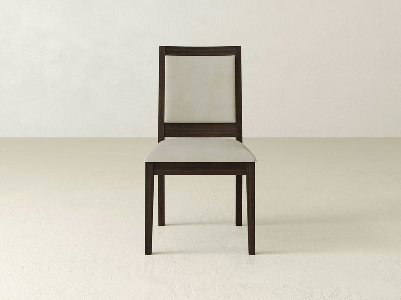 The Gina Side Dining Chair - Tobacco, a product by James+James, is centered in an empty, neutral-toned room with a plain wall and floor. It features a wooden frame with a light-colored upholstered backrest and seat. The chair boasts a simple and minimalist design with clean lines.