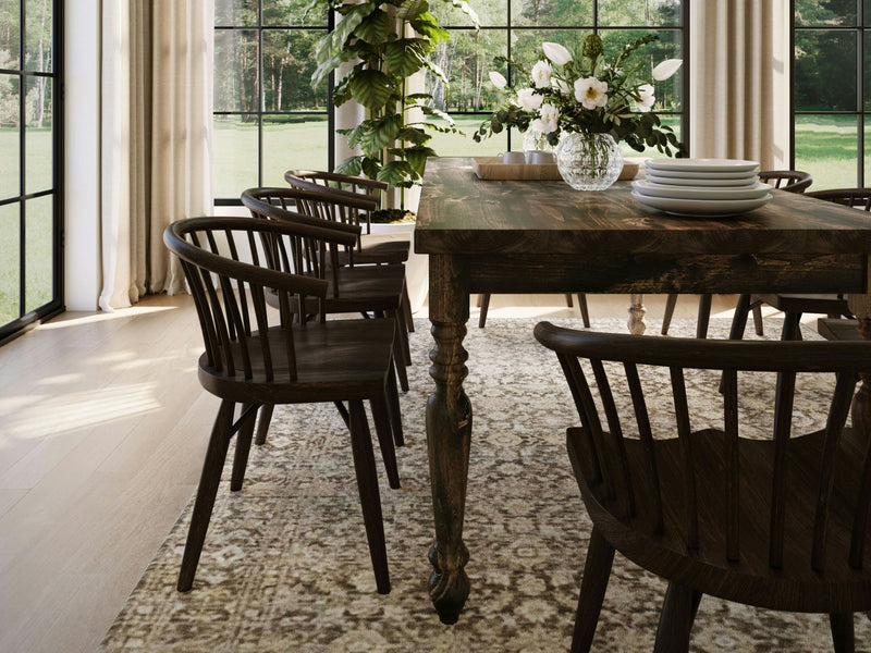 A dining room featuring the French Country Dining Table in Tobacco by James+James is surrounded by dark wooden chairs. The table is elegantly set with a stack of plates and a flower arrangement in a glass vase. Large windows in the background overlook a lush green outdoor area, allowing abundant natural light to flood the space.