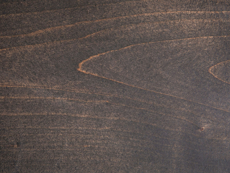 A close-up of the Deep Grey on Alder Sample from James+James showcases a dark, rich brown hue with prominent grain patterns. The texture reveals natural lines and swirls, imparting an organic and rustic appearance.