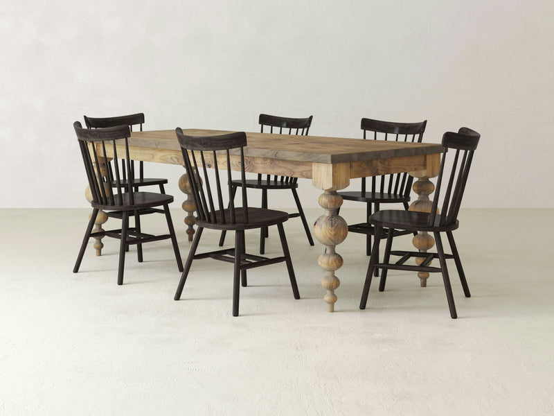 The Olivia & Rustic Windsor Dining Set by James & James features an Olivia dining table with ornate, turned legs, surrounded by six dark wooden rustic Windsor dining chairs with spindled backs. The set rests on neutral-colored, textured flooring against a light wall, evoking a rustic and minimalist aesthetic.