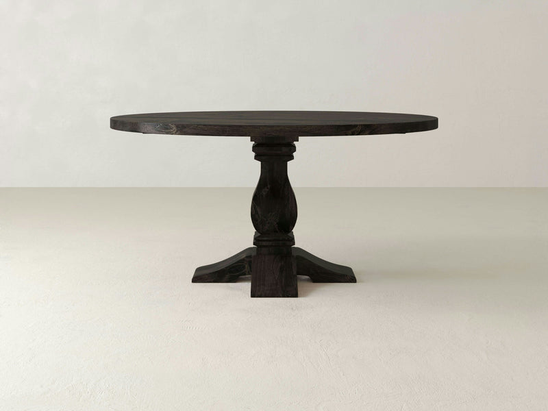 The Heirloom Round Dining Table - Charred Ember by James+James features a smooth, dark wooden surface with a classic design. It stands on a single pedestal leg with four feet and is set against a light-colored background, offering a simple yet elegant appearance.