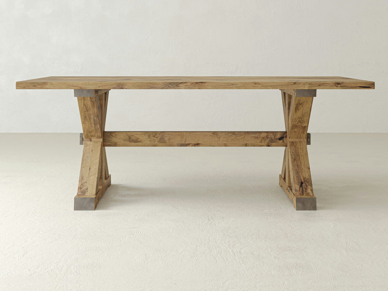 A large Trestle Dining Table - Harvest Wheat from James+James stands on a plain, off-white floor against a light gray wall. The table's surface and legs exhibit a natural wood grain, showcasing a rustic and sturdy design, while the trestle base features metal accents at the joints.