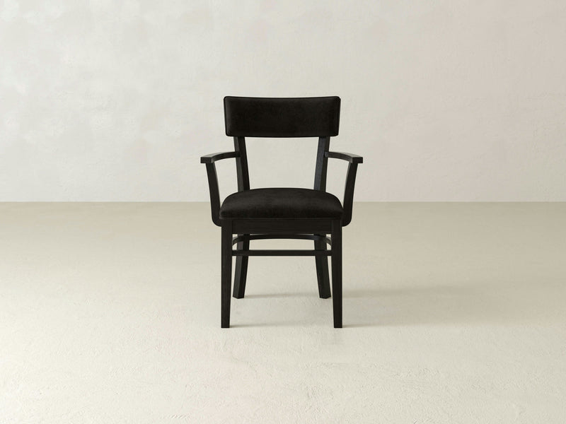 The Everly Arm Dining Chair - Charred Ember from James+James is centered in the image. This black wooden chair features a cushioned backrest and seat, along with armrests, and is placed on a light-colored floor against a plain, light-colored background.