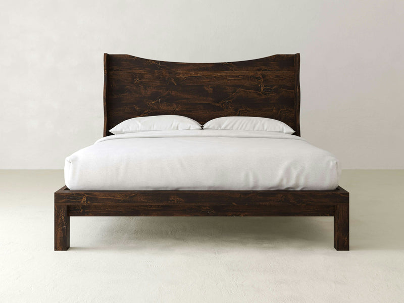 In a minimalist room, the Harlow Bed - Tobacco by James+James stands out with its unique, angular headboard design and dark wooden frame. The bed is neatly made with crisp white bedding, including two pillows. The plain, light-colored background wall and floor accentuate the bed as the focal point.