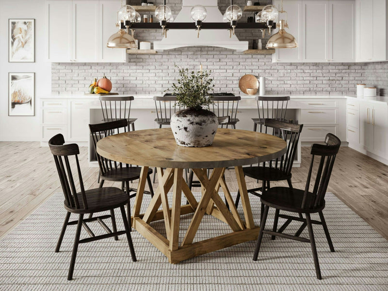 A modern kitchen features the Julia Round Dining Table in Harvest Wheat from James+James with a black and white potted plant centerpiece, surrounded by six black chairs. In the background, white cabinets, a brick backsplash, three pendant lights, and a range hood are visible. The floor has a textured rug.