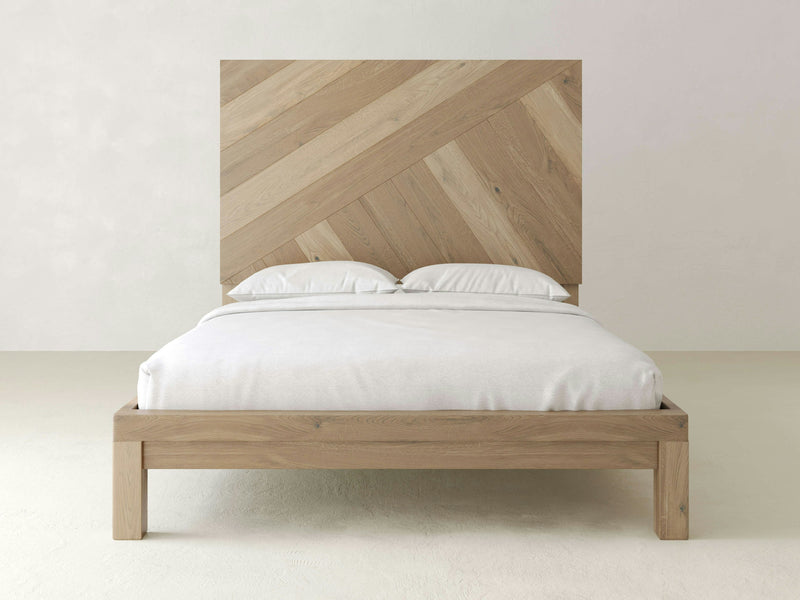 The Luna - Barn Wood bed by James+James features a minimalist design with a large, patterned headboard and clean, simple lines. Made from light-colored wood and dressed in white linens, it sits against a plain white wall in a bright, spacious room.