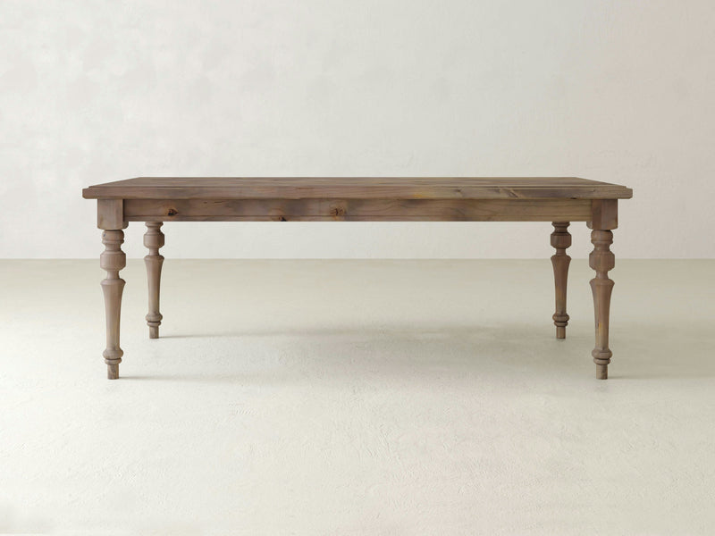 The Allyson Dining Table by James+James, featuring a rectangular top and four intricately carved legs, is placed against a simple, light-colored background. This dining table boasts a rustic finish that highlights the natural grain and texture of the barn wood.