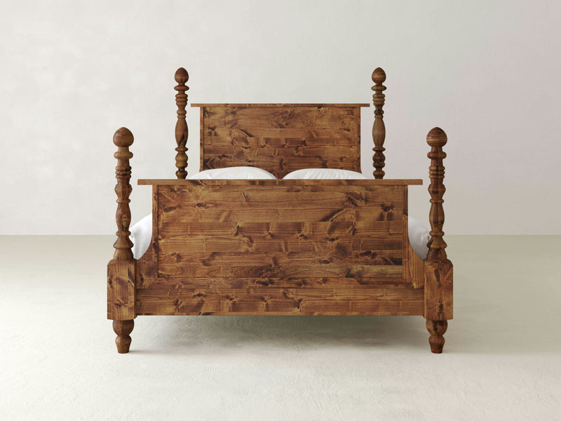 The Catherine Bed - Tuscany by James+James is a wooden bed with a rustic design, featuring intricately carved posts at each corner and a high headboard and footboard. Set against a plain, light-colored background, the wood grain and texture are prominently visible, showcasing detailed craftsmanship.