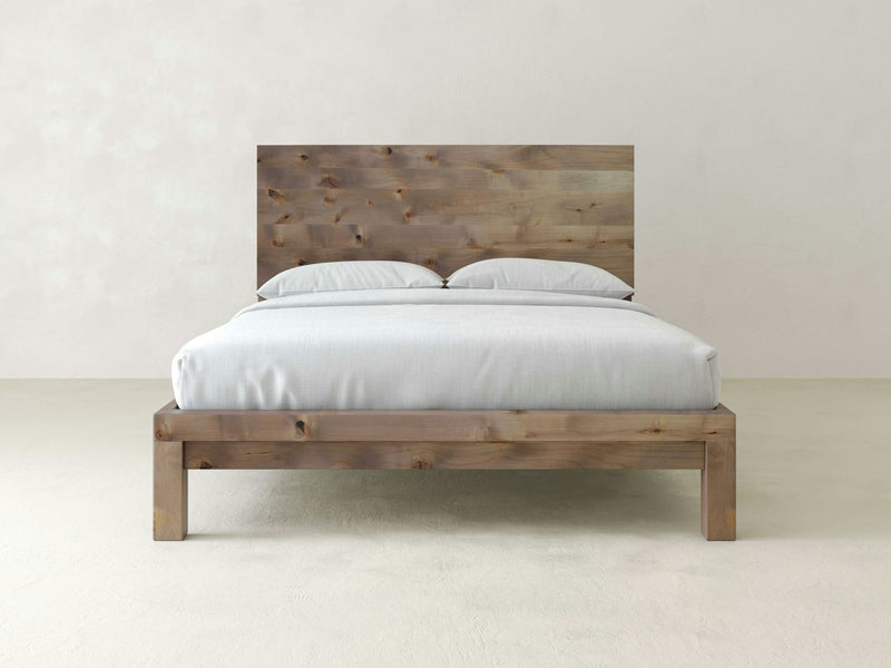 A neatly made Sawyer Bed - Barn Wood by James+James, featuring a wooden frame and headboard, stands against a plain, light-colored wall. The bed is dressed in white linens and two pillows, showcasing a minimalist and modern design.