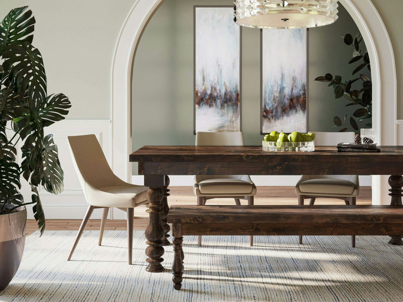 A well-lit dining area features a rustic wooden table accompanied by the Annli Bench - Tobacco from James+James, along with beige upholstered chairs. A glass bowl filled with green apples sits atop the table. In the background, two abstract paintings and a potted plant add to the decor. Above the table hangs a chandelier, casting a warm glow over the setting.