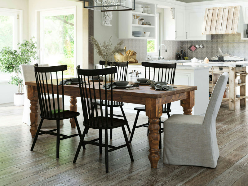 A cozy kitchen featuring the Baluster Dining Table - Tuscany by James+James, surrounded by black and upholstered chairs. The white cabinets and light-colored walls create a bright atmosphere. Decorative items adorn the shelves, a potted plant sits near the window, and kitchen utensils are neatly arranged on the countertop.