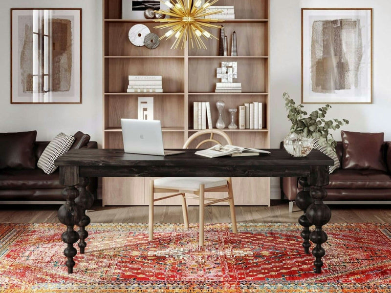 A stylish home office is anchored by the Olivia Desk in Charred Ember from James+James, featuring ornate legs. On the desk sit a laptop and an open book. Behind the desk is a light wooden chair, flanked by two leather armchairs. The room's look is completed with a bookshelf adorned with decor objects and a large modern chandelier.