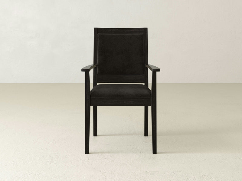 The Gina Arm Dining Chair by James+James boasts a contemporary silhouette, crafted from wood with dark-colored upholstery set against a plain, light-colored backdrop. Featuring armrests and a straight, rectangular backrest, this chair's minimalist design exudes modern elegance.