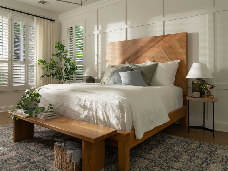 A cozy bedroom showcases a large wooden Luna - Satin bed by James+James, adorned with white bedding and multiple pillows. Two bedside tables with lamps flank the bed. A bench sits at the foot, with a wicker basket beneath. Sunlight streams in through plantation shutters, highlighting the green plants.