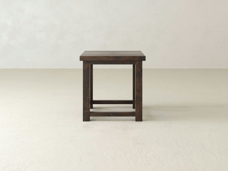 The Carter X Side Table - Tobacco by James+James is centered against a plain, light-colored wall and floor. This dark wooden table with a rectangular top features a minimalist design and includes sturdy, straight legs connected by a horizontal support bar at the bottom.