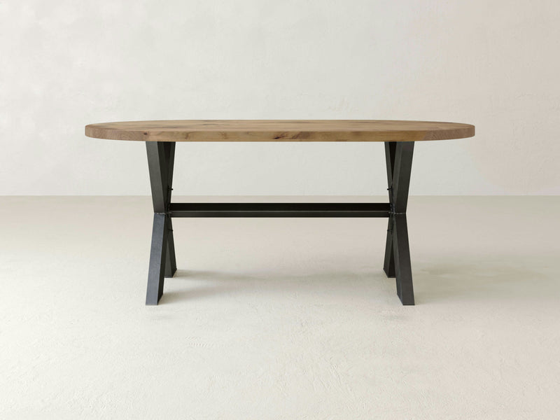 The X-Base Oval Dining Table - Harvest Wheat by James+James features a simple, oval-shaped wooden top with a light brown finish. It has black, metal trestle-style legs with an underside crossbar for support. The table is set against a plain, light-colored background.