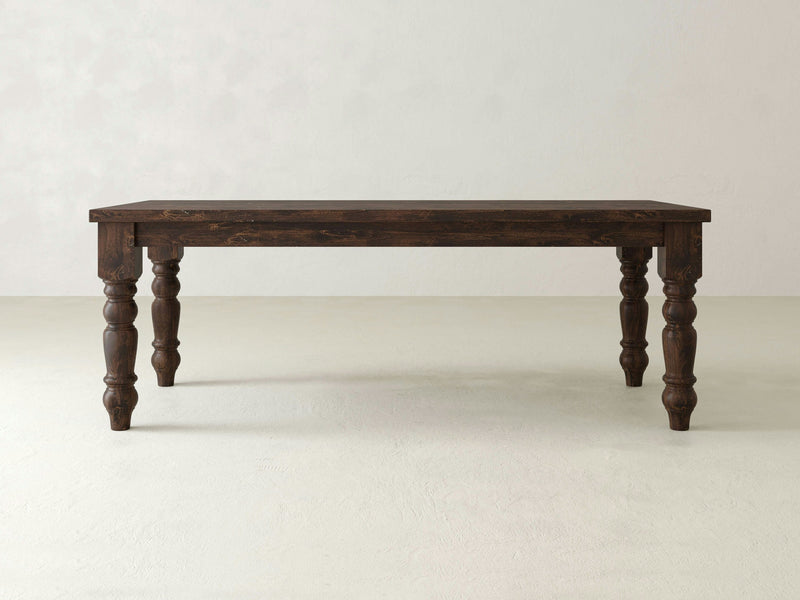 The Baluster Dining Table - Tobacco by James+James is a dark brown wooden dining table with an ornate design, featuring four intricately carved legs and a rectangular top. The background is a minimalist, light-colored wall and floor.