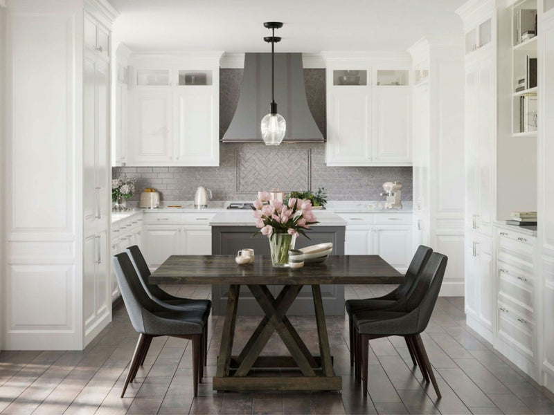 A modern kitchen featuring white cabinets, a stainless steel range hood, and a herringbone backsplash. Centered in the dining area is the Julia Square Dining Table - Charred Ember from James+James, paired with four black chairs. The table is adorned with a vase of pink flowers, contributing to the room's well-lit, airy feel.