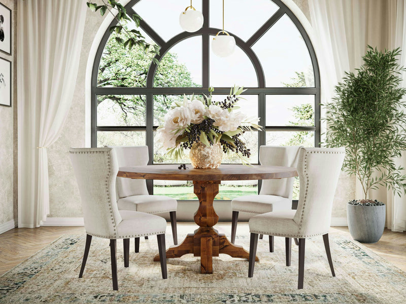 A stylish dining room featuring the Sybil Round Dining Table - Tuscany by James+James, surrounded by four upholstered chairs and placed on a patterned rug. The table is adorned with a floral centerpiece. Large arched windows in the background let in natural light, with lush greenery visible outside. White curtains frame the windows.

