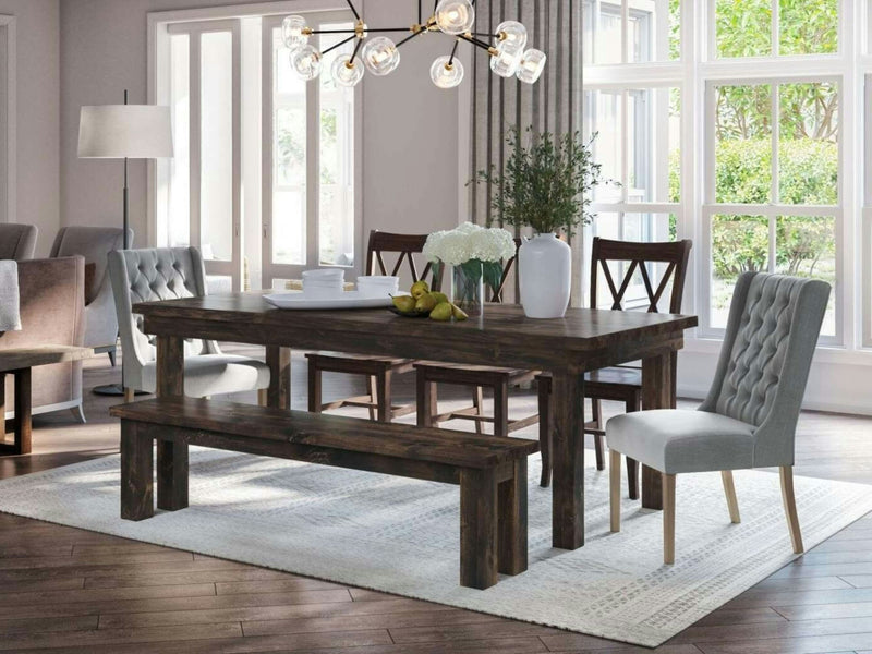 In a well-lit dining room, the Farmhouse Expandable Dining Table - Tobacco by James+James takes center stage with its rustic wooden design. A vase of white flowers and a bowl of fruit adorn the table, which is surrounded by four wooden chairs with cushioned seats and one wooden bench. Above, a modern chandelier provides illumination, while large windows allow natural light to flood the space.