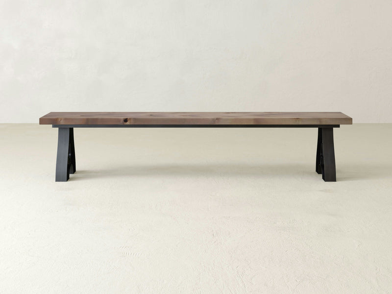 The Wishbone Bench - Barn Wood by James+James, featuring a minimalist design with a rectangular, flat seat and black, angled legs, stands on a light-colored floor against a plain, off-white wall. Its simple and rustic design showcases the natural wood grain patterns on the seat.