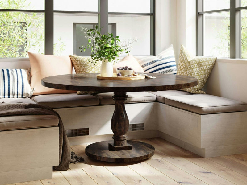 A cozy dining nook featuring the Vivien Round Dining Table in Tobacco by James+James, with a vase of green foliage at its center. The built-in bench seating is adorned with various patterned cushions and pillows, bathed in natural light from the surrounding windows.