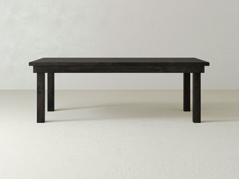 A sleek, rectangular wooden table with a charred ember finish. The Farmhouse Dining Table by James+James features four straight legs and a smooth top surface. It is set against a plain, textured light-colored wall and sits on a light-colored floor.