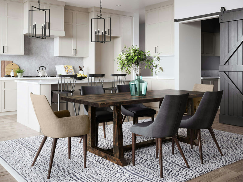 A modern dining area showcases the Cora Dining Table in Tobacco by James+James, surrounded by a mix of beige and gray chairs on a patterned blue and white rug. Behind the table, a kitchen area with white cabinets and a central island featuring black stools is visible. A sliding barn door is on the right.