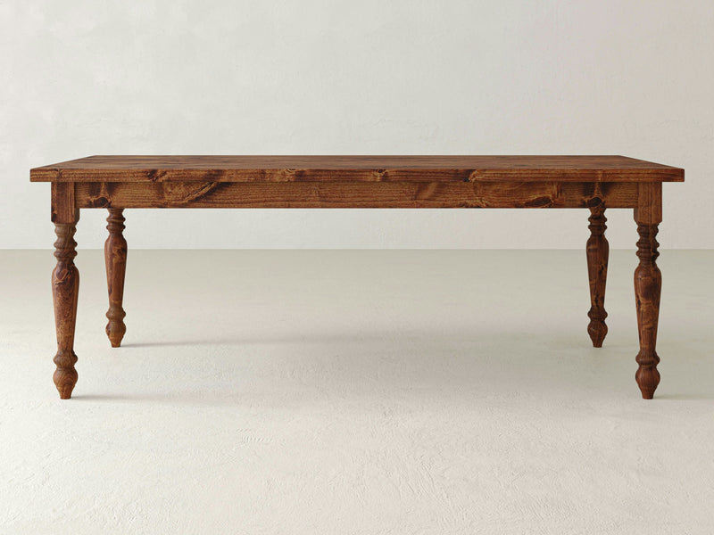 A James+James French Country Dining Table - Tuscany, crafted with sturdy wood and a rich brown finish, featuring intricately carved legs and a smooth rectangular top. The table is set against a plain, light-colored background.