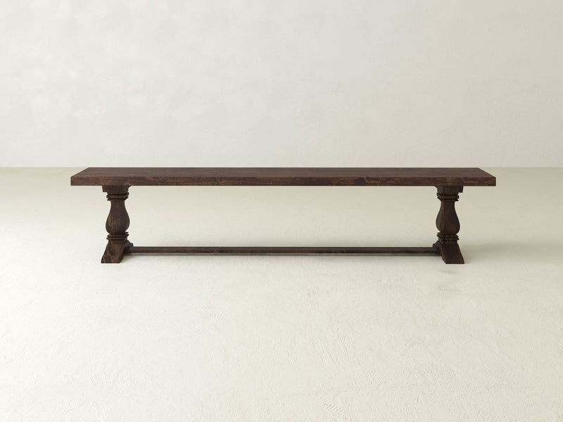 Introducing the Heirloom Bench by James+James: a long, rectangular wooden masterpiece with a dark brown finish. This elegant bench features two meticulously carved legs on each end, connected by a horizontal support bar, exuding timeless sophistication against a plain, light-colored background.