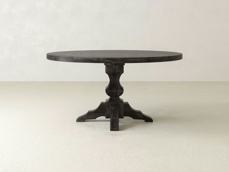 A Sybil Round Dining Table - Charred Ember by James+James stands against an off-white, minimalist background. This dark wooden round table features a smooth surface and a sturdy central support with carved detailing, elegantly supported by a simple pedestal base.