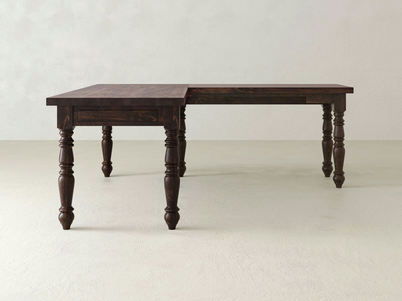 The Turned Leg Corner Desk - Tobacco from James+James is displayed in a light grey room. The dark wooden desk features a rectangular shape with decorative, turned legs and boasts a rich, polished finish on both the tabletop and legs. It is positioned to appear as if extending, with the corner section partially pulled out.
