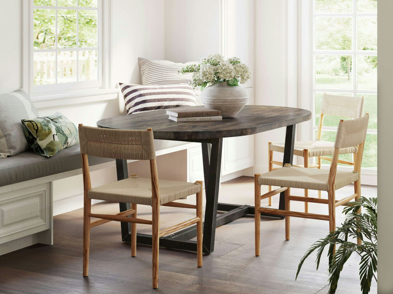 A cozy dining nook features the Trapezoid Oval Dining Table in Tobacco by James+James, surrounded by three beige woven chairs. A gray cushioned bench with patterned throw pillows lines the window, and a vase with white flowers sits on the table. Large windows let in abundant natural light.