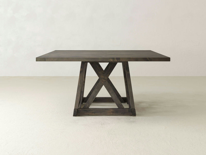 The Julia Square Dining Table - Charred Ember by James+James is a square wooden dining table with a dark brown finish. It has a sturdy trestle base featuring an "X" design on each side for structural support. The table is positioned against a plain, light-colored background.