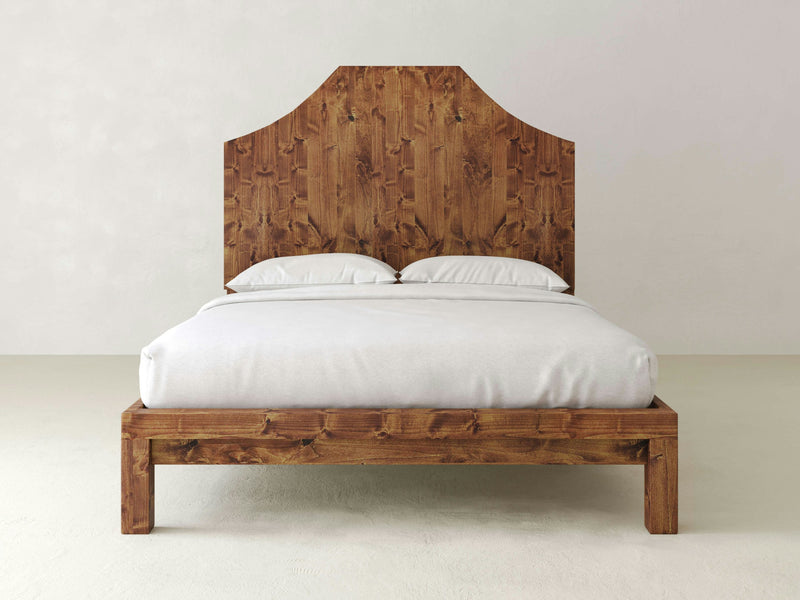 Introducing the Megan Bed - Tuscany from James+James: a wooden bed frame with a tall, intricately shaped headboard, featuring a rustic, natural wood finish. The bed is perfectly made with white bedding and two white pillows against a plain, light-colored background.