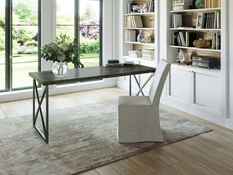 A modern, bright home office with a large window offering a garden view. The room features the Luca Desk in Deep Grey from James+James, characterized by its sleek wooden top and sturdy metal legs. A cushioned chair accompanies the desk, while a decorative vase with flowers adds a touch of elegance. Built-in bookshelves are filled with books and decor items, and a light area rug covers the floor for added comfort.