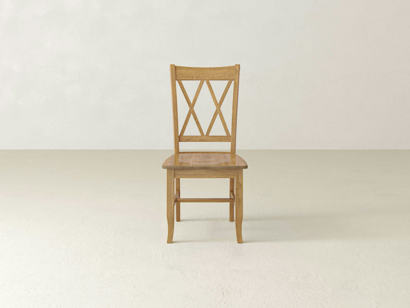 A single Double X-Back Dining Chair - Harvest Wheat by James+James is centrally placed against a plain, light-colored background. The chair has a rectangular seat and a backrest featuring a double-crossed design. The floor and wall are both light beige.