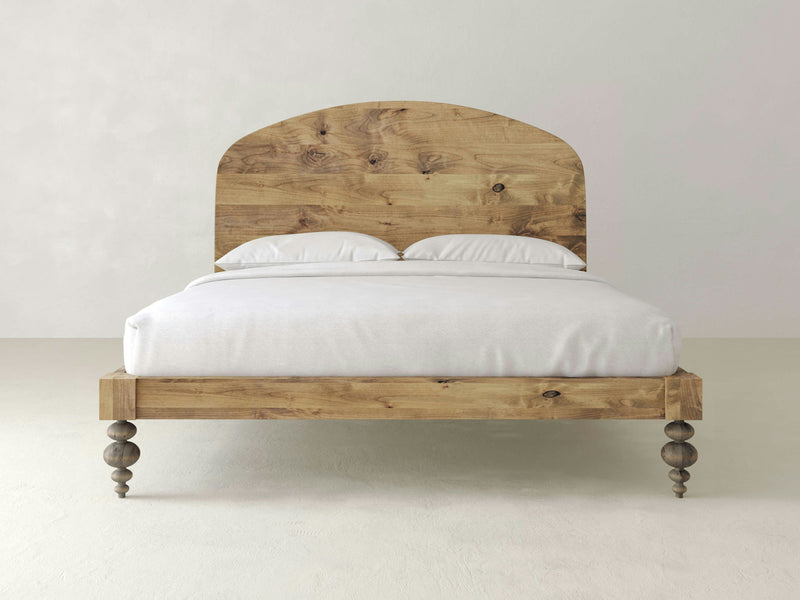 The Eden Bed - Harvest Wheat from James+James is a wooden bed featuring a natural finish and a curved headboard. It is adorned with a white duvet and two white pillows, and stands on turned wooden legs against a plain white background.