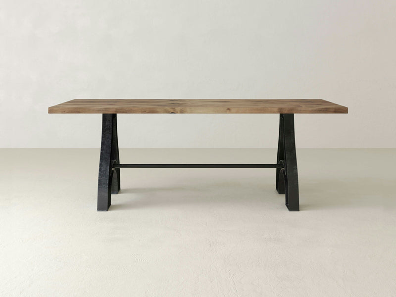 The Wishbone Dining Table - Harvest Wheat by James+James features a rectangular wooden top and sleek black metal legs in an A-frame design. The table showcases a minimalist and modern aesthetic, standing on a neutral light-colored floor against a plain white wall.