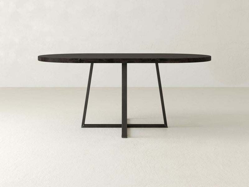 A Watson Oval Dining Table - Charred Ember by James+James is centered in the image. It features a modern, minimalist design with a dark wooden finish and a thin metal base forming a geometric support structure. The background is a plain, light-colored wall and floor, accentuating the table’s sleek appearance.