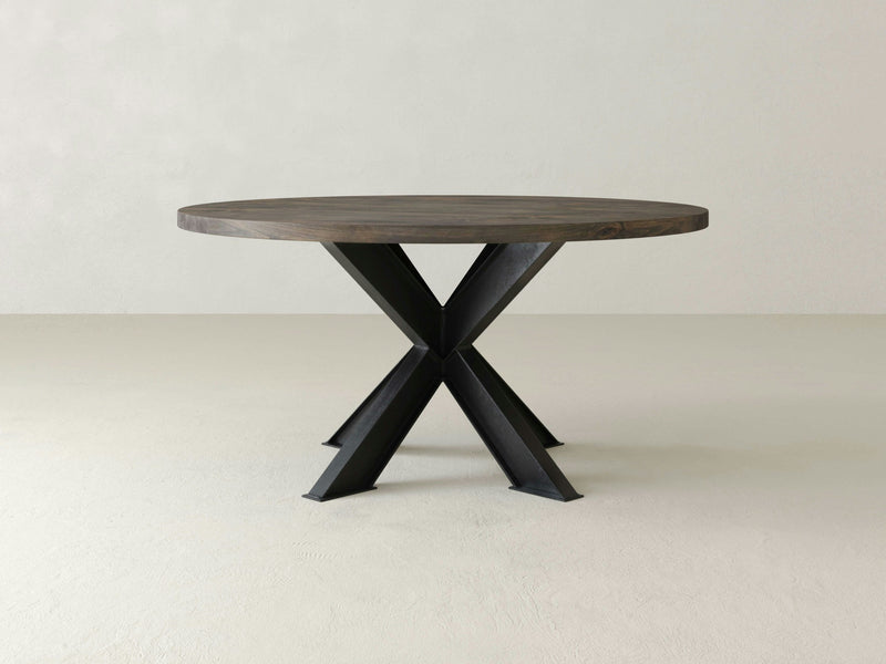 The Shiloh Round Dining Table – Deep Grey by James+James features a round wooden top with a dark finish, supported by an X-shaped black metal base. The table is centered and placed against a plain, light-colored background.