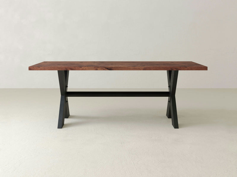 A rectangular X-Base Dining Table - Tuscany by James+James, featuring a dark brown wooden top and black metal X-shaped legs, stands on a plain, light-colored floor against a neutral background.