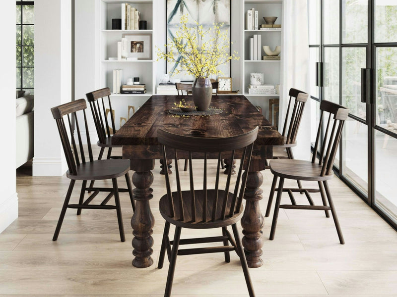 A rustic dining room featuring the Annli Dining Table in Tobacco from James+James with six matching chairs. On the table, a vase with yellow flowers serves as a centerpiece. In the background, built-in white shelves filled with books and decor, and large glass windows let in natural light.